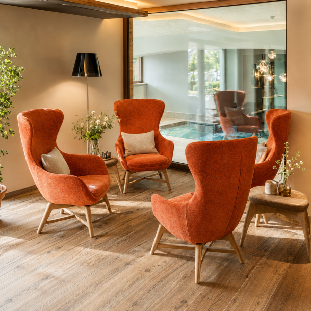Area relax di centro benessere con poltrone imbottite di design, pavimento effetto legno e vista sulla piscina interna attraverso una grande vetrata.
