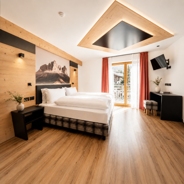 Camera d’hotel in stile alpino contemporaneo con arredi su misura in legno, letto matrimoniale, pavimento effetto legno e balcone panoramico.