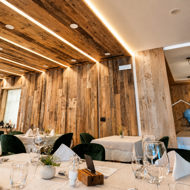 Dettaglio della sala ristorante dell'Hotel Olimpionico con pareti e soffitto in legno anticato, illuminazione LED integrata, tavoli apparecchiati e sedute verdi dal design contemporaneo realizzati da Arredamenti Depal.