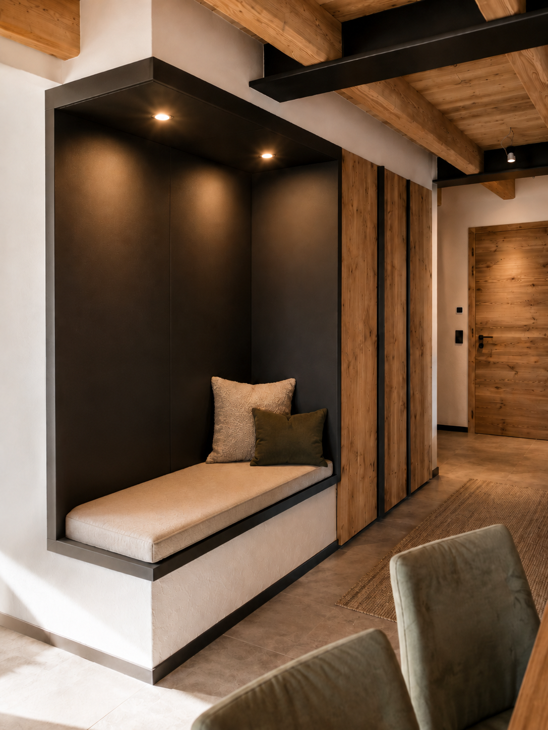 Panca su misura integrata in una nicchia rivestita in finitura scura, illuminata da faretti incassati e completata da morbidi cuscini decorativi. Sullo sfondo, armadiature in legno e dettagli neri definiscono un elegante ingresso in stile alpino contemporaneo.