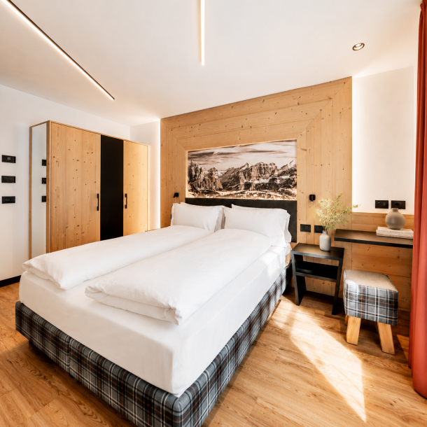 Camera d’hotel in stile alpino moderno con arredi su misura in legno, testiera decorativa con panorama montano, scrivania integrata e illuminazione contemporanea.
