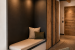 Panca su misura integrata in una nicchia rivestita in finitura scura, illuminata da faretti incassati e completata da morbidi cuscini decorativi. Sullo sfondo, armadiature in legno e dettagli neri definiscono un elegante ingresso in stile alpino contemporaneo.