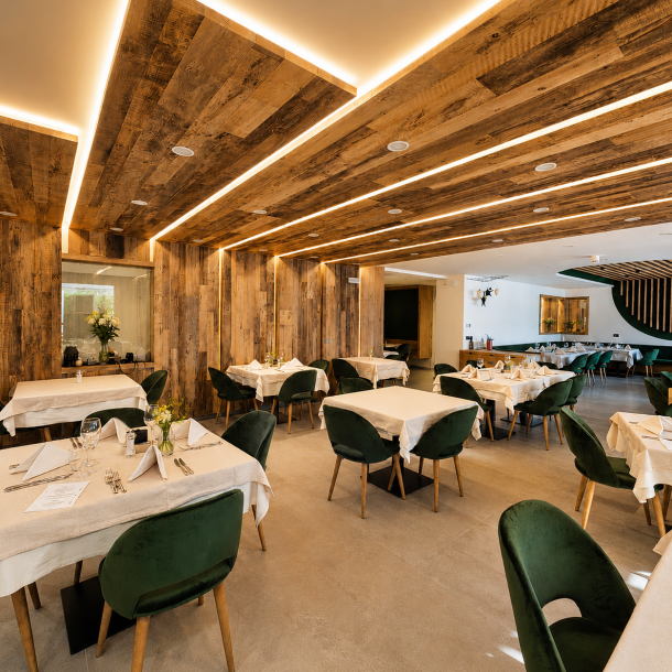 Sala ristorante dell'Hotel Olimpionico con rivestimenti in legno antico, illuminazione integrata a LED, tavoli apparecchiati e sedute verdi in stile alpino contemporaneo realizzata da Arredamenti Depal.