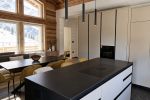Cucina moderna su misura per chalet di montagna con grande isola centrale in finitura nera e bianca, colonne attrezzate a tutta altezza e soffitto in legno a vista. Le ampie vetrate incorniciano il panorama alpino innevato, mentre il design essenziale crea un raffinato equilibrio tra tradizione e contemporaneità.
