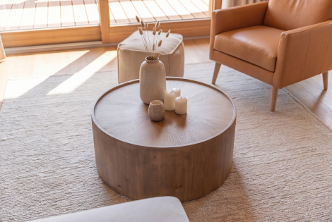 Tavolino da salotto rotondo in legno naturale con finitura materica, decorato con elementi in ceramica e dettagli botanici. Un complemento d'arredo elegante inserito in un ambiente luminoso con poltrona in pelle e ampie vetrate.